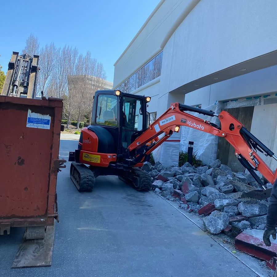 Compact excavator breaking a concrete entry beside a roll-off container