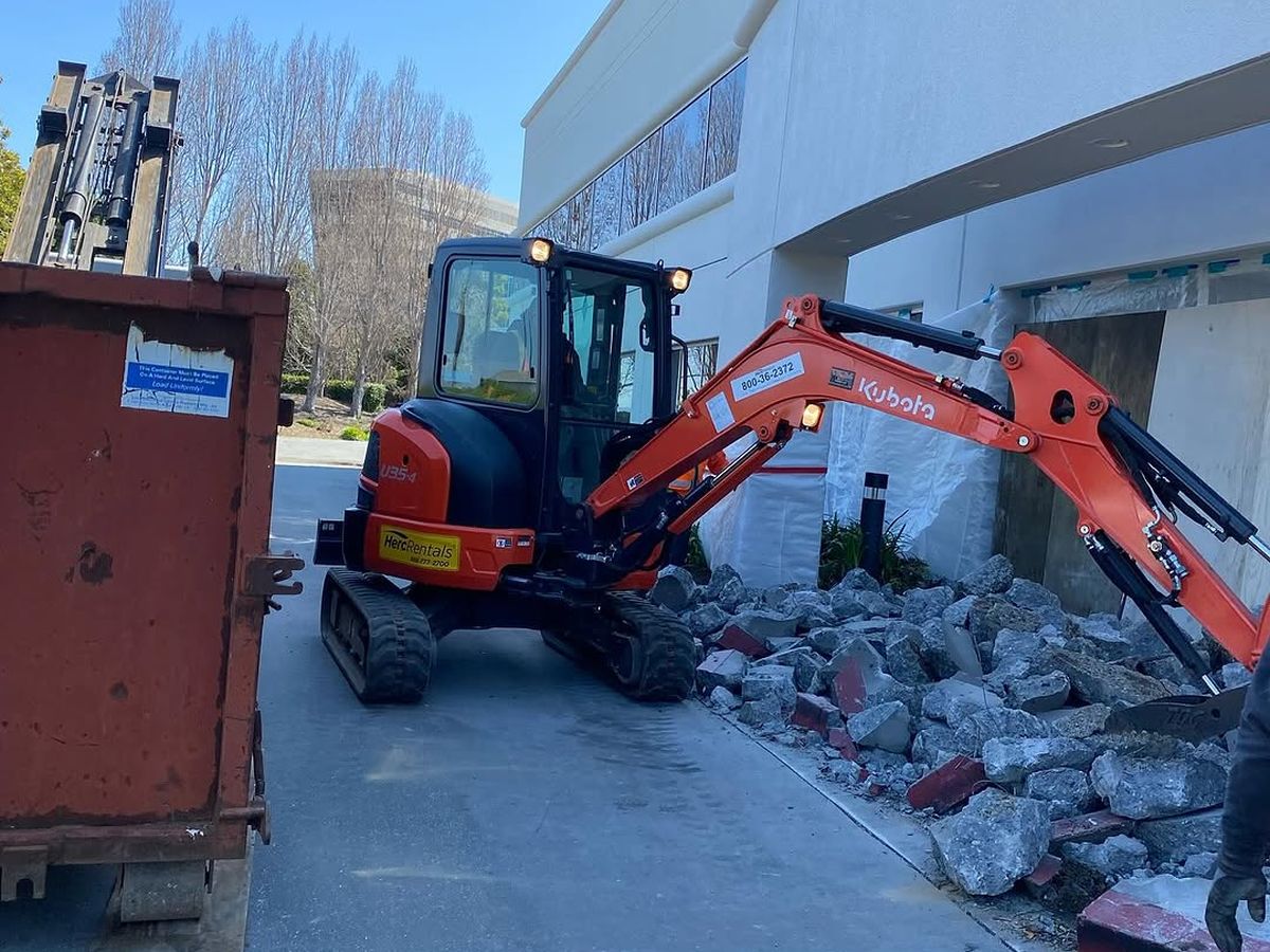 Compact excavator breaking out a concrete entry slab next to a waiting roll-off container