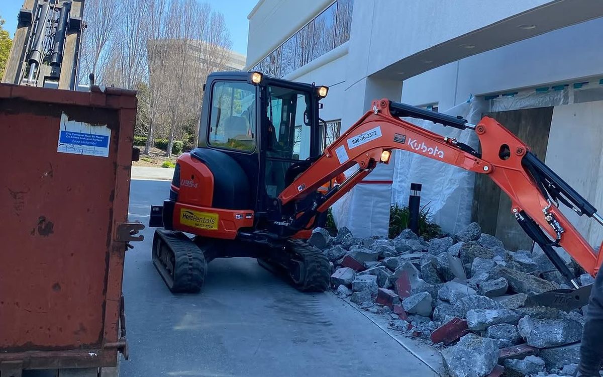 Compact excavator breaking a concrete entry into pieces beside an open container
